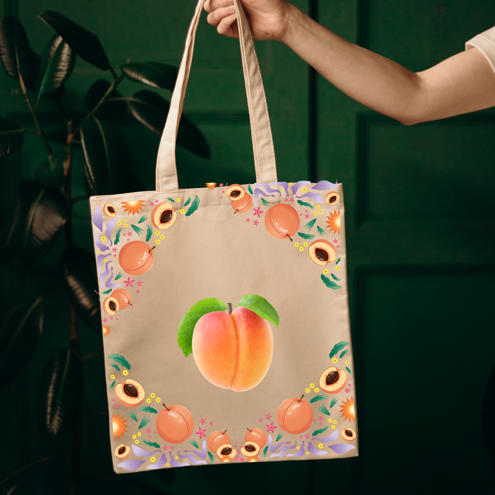 A canvas tote bag with a peach illustration and floral border, held by a person's hand against a dark green background.
