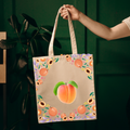 A canvas tote bag with a peach illustration and floral border, held by a person's hand against a dark green background.