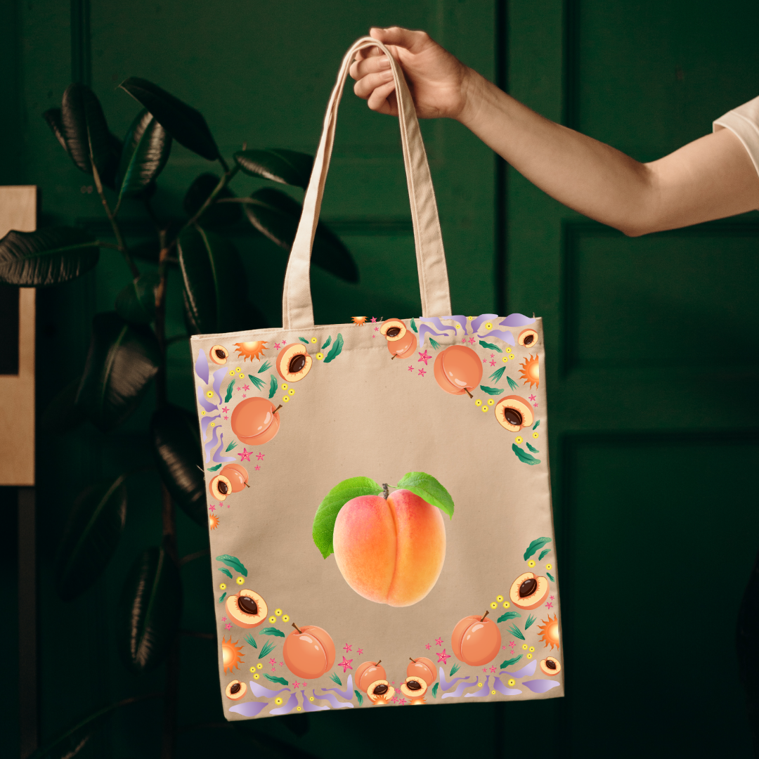 A canvas tote bag with a peach illustration and floral border, held by a person's hand against a dark green background.