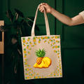 A beige canvas tote bag with a colorful pineapple print, held by a person's arm against a dark green background.