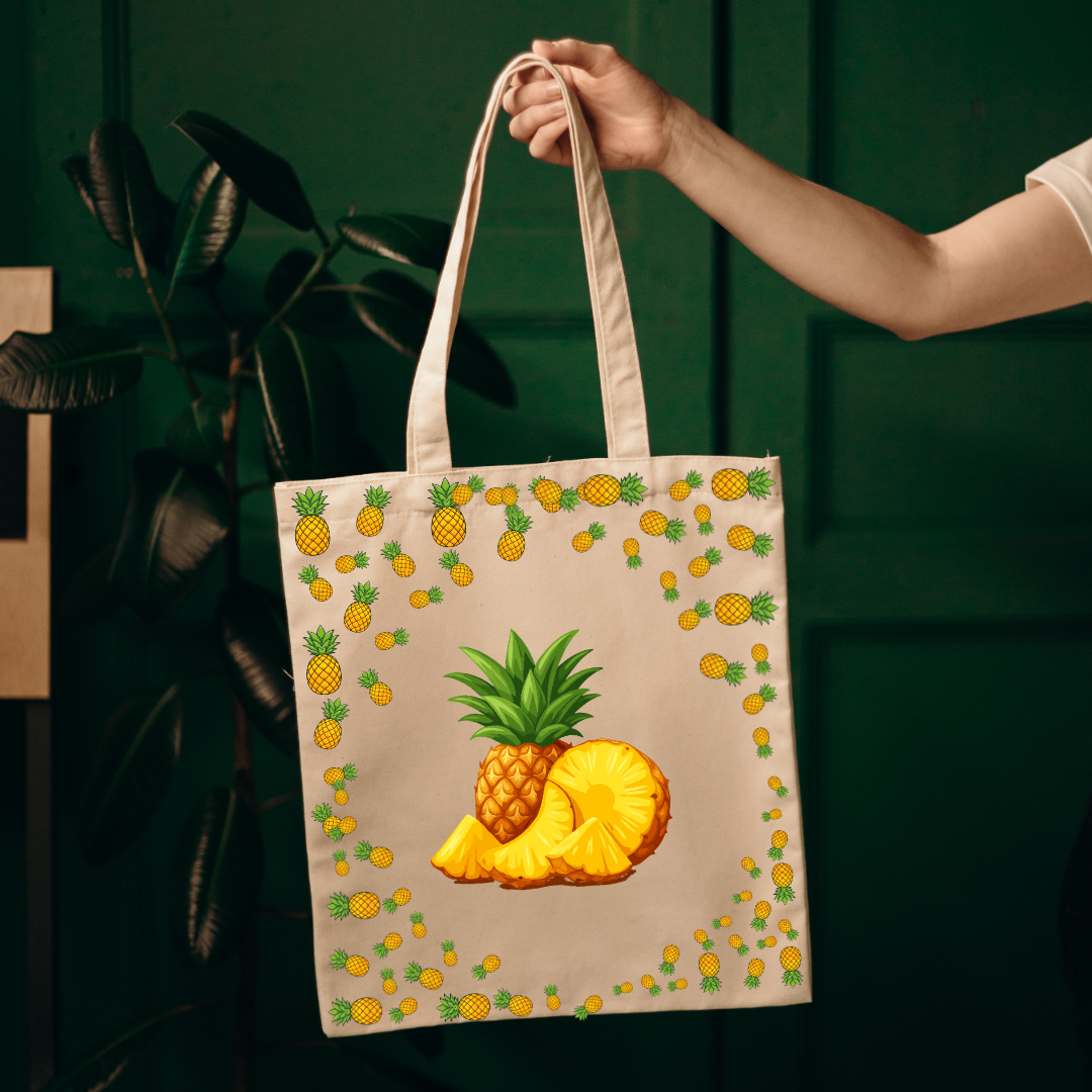 A beige canvas tote bag with a colorful pineapple print, held by a person's arm against a dark green background.