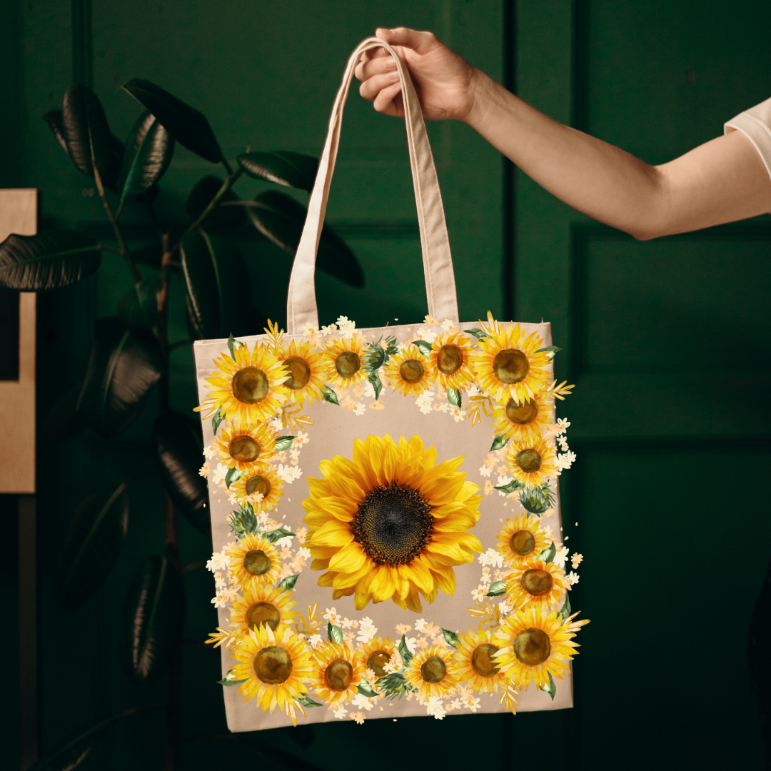 A canvas tote bag with a sunflower print, held by a person's hand against a dark green background with a plant.