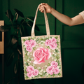A beige canvas tote bag with a large pink rose and surrounding pink roses and green leaves, held by a person's hand against a dark green paneled background.