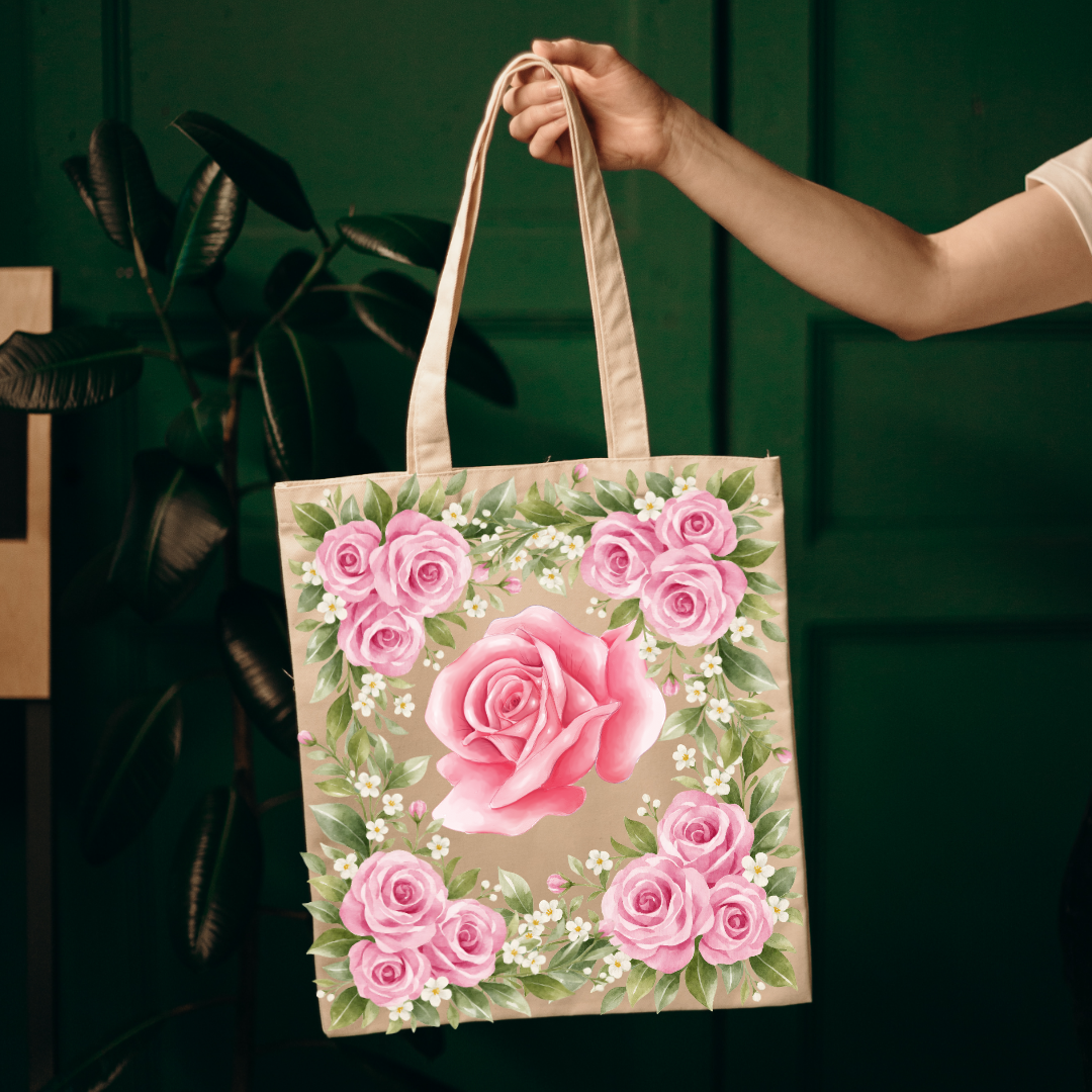 A beige canvas tote bag with a large pink rose and surrounding pink roses and green leaves, held by a person's hand against a dark green paneled background.